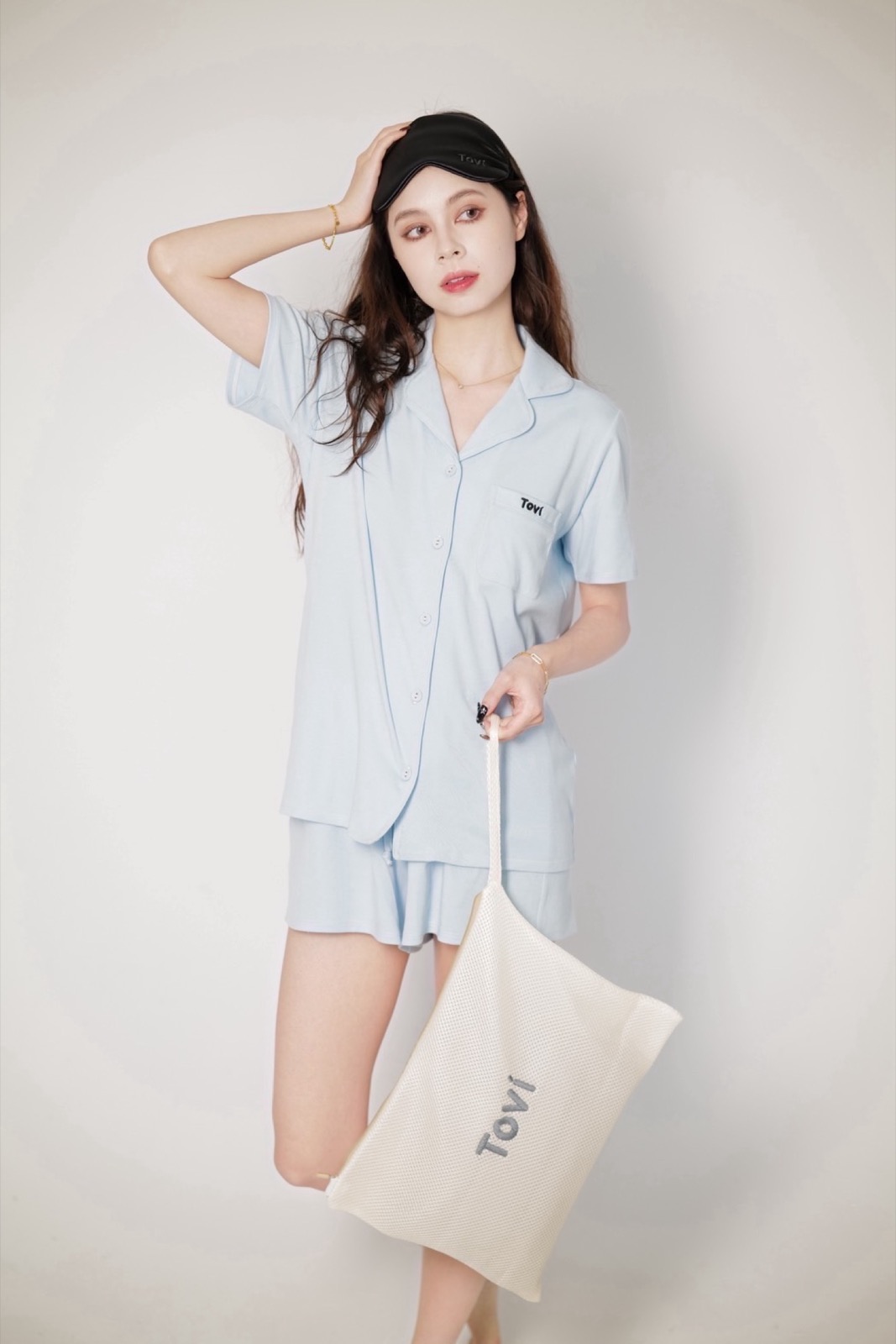 Model in pale blue Tovi sleepwear holding a cream travel pouch and wearing a sleep mask